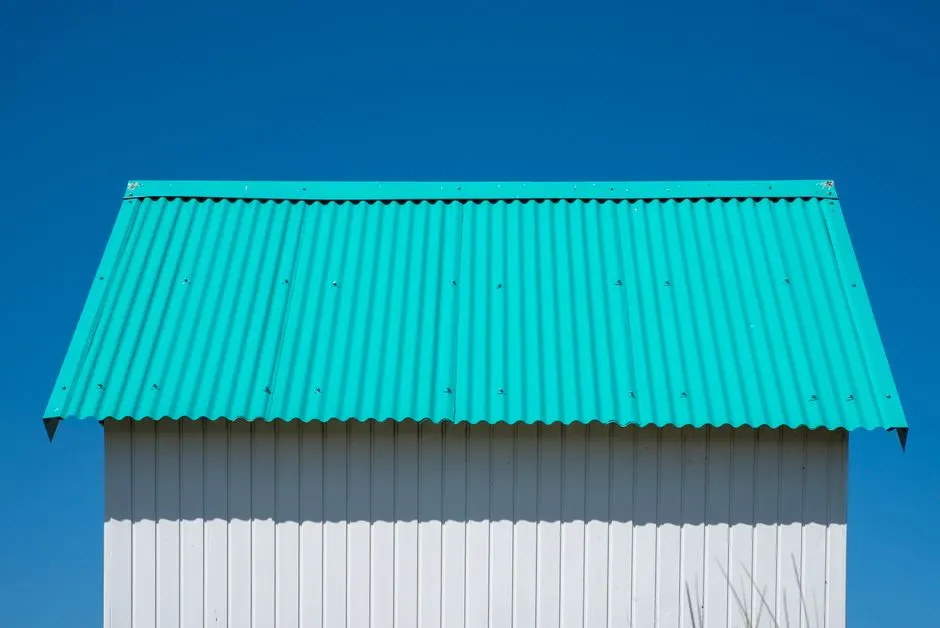 Simple architectural composition with a teal corrugated roof and white wall under clear blue sky.